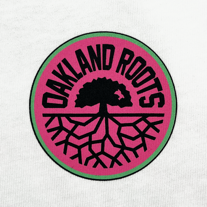 Check out the Staycation Heavy Tee by Oakland Roots SC, featuring a circular logo with a green border and a magenta background. At the center, a black silhouette of a tree spreads its roots like branches into the lower half of the circle. Bold, black letters arch above reading "OAKLAND ROOTS," emphasizing social good and community connection.
