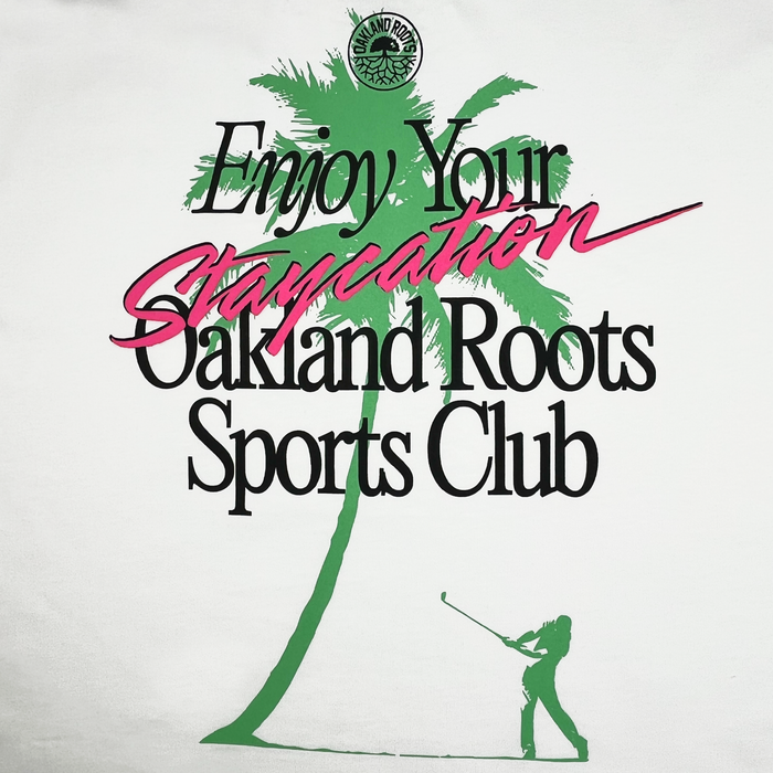 A graphic showcases a green silhouette of a palm tree and a person swinging a golf club, with overlay text that reads "Enjoy Your Staycation" in cursive, and "Staycation" highlighted in pink. Below it says "Oakland Roots SC" in black. The small circular logo appears in the upper left corner, making it perfect for the Oaklandish Boxy fit Staycation Heavy Tee from Oakland Roots Sports Club promoting social good.