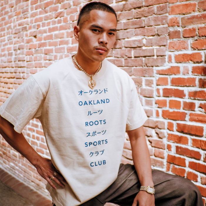 A man poses confidently by a red brick wall, wearing the Oakland Roots SC Story Telling Tee featuring blue Japanese and English text. He pairs it with jewelry and brown pants, striking a stylish stance with one hand on his hip.
