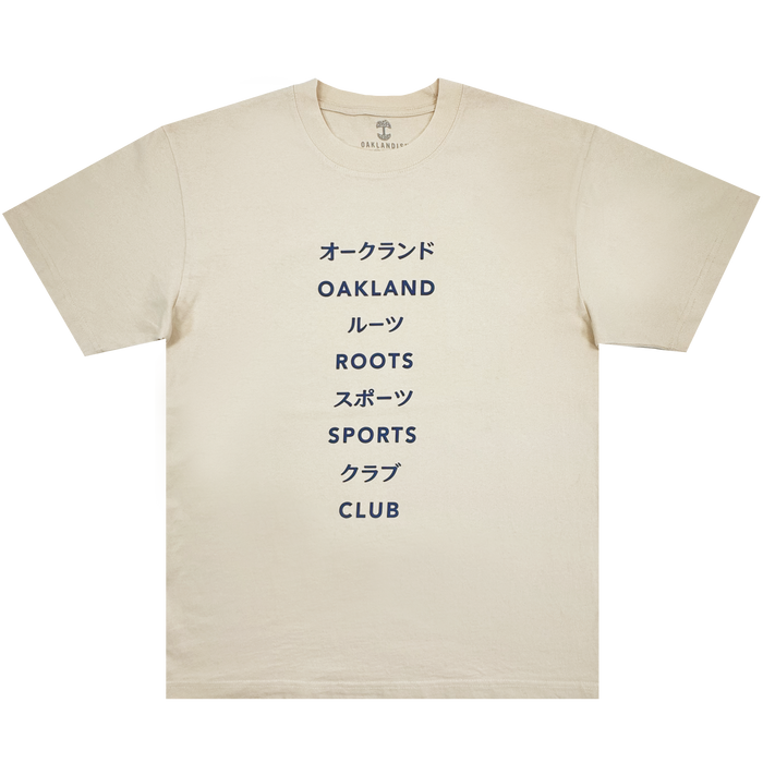 The Story Telling Tee by Oakland Roots SC is a cream tee featuring bold navy text, with alternating Japanese katakana and English on the chest: “オークランド OAKLAND ルーツ ROOTS SC スポーツ SPORTS クラブ CLUB.”.