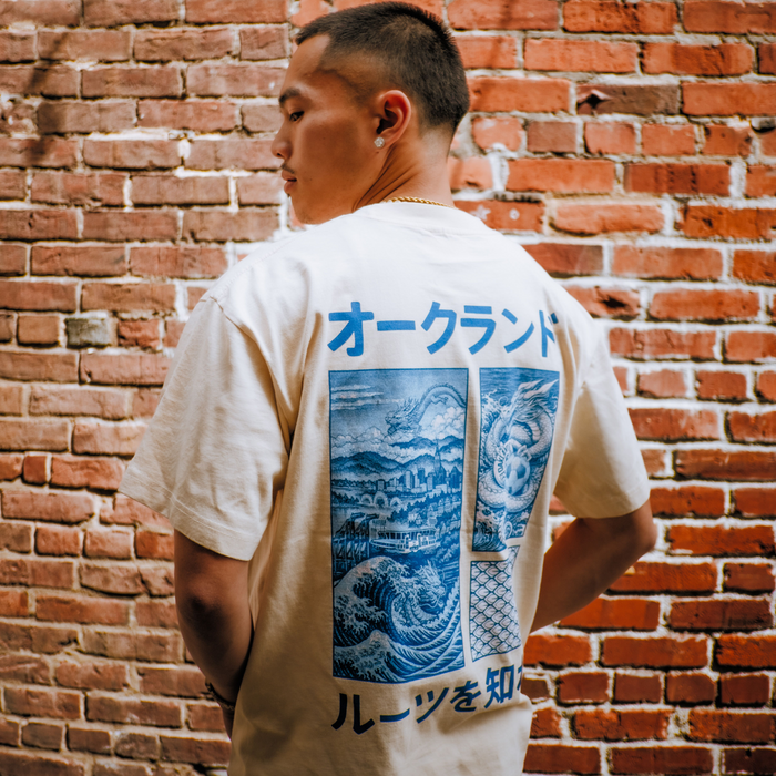 A young man with short hair and a chain necklace stands sideways by a brick wall, wearing the cream Story Telling Tee from Oakland Roots SC featuring blue Japanese text and ocean wave art on the back, looking to the side seriously.
