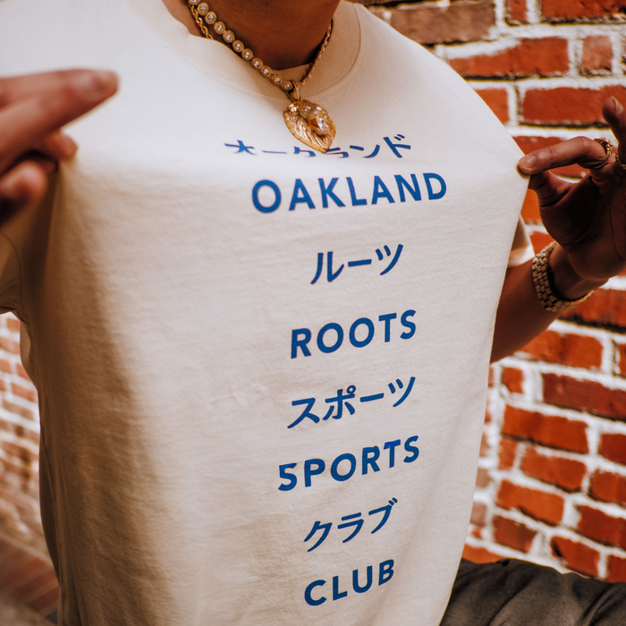 Wearing the Story Telling Tee by Oakland Roots SC, featuring “OAKLAND ROOTS SPORTS CLUB” in English and Japanese, a person poses with gold jewelry against a red brick wall, showcasing their Roots SC style.