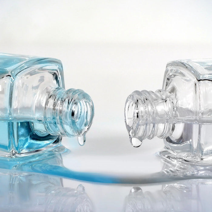 Two LONDONTOWN Strength & Shine Duo bottles spill vibrant blue nail strengthener onto a reflective surface, showcasing the secret to shiny, strong nails.