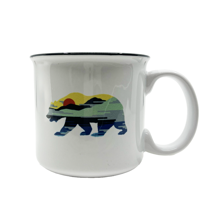The Sunset Bear Coffee Mug by Oaklandish is a white ceramic mug with a thick black rim, showcasing a graphic of a California grizzly bear filled with a landscape design. The design features a serene outdoor scene with layers of hills and water bathed in the colors of a colorful sunset, including shades of green, yellow, orange, and blue.
