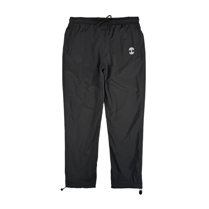 The Oaklandish Susurrus Track Bottom is a pair of black nylon pants featuring an adjustable waistband, drawstring cuffs, two side pockets, and a small white circular logo on the left thigh. Set against a plain white background.
