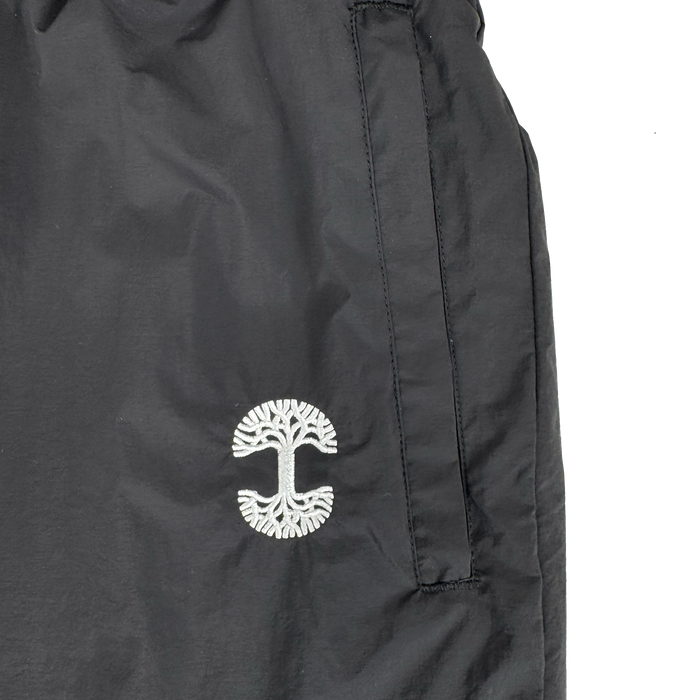 Close-up of the Oaklandish Susurrus Track Bottom in black nylon, featuring a white embroidered tree logo near the pocket, vertical zipper pocket opening, and visible stitching details. Set against a plain white background to highlight its modern style.