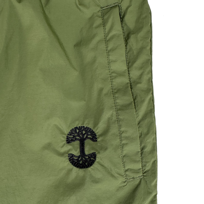 Close-up of Oaklandish's Susurrus Track Bottom in olive green nylon, with a slanted pocket and black tree logo below it. The plain white background highlights its color, texture, and adjustable waistband.