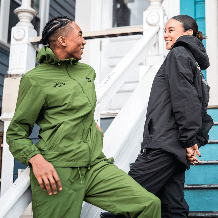 Two people smile at each other on a porch. The person on the left wears green nylon pants with an adjustable waistband. The person on the right stands by a white door in Oaklandish Susurrus Track Bottoms, featuring an embroidered tree logo.