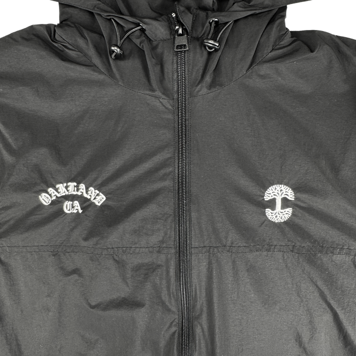 The Susurrus Track Top by Oaklandish is a black nylon zip-up jacket with a subtle sheen. "OAKLAND CA" is printed in white Old English on the left chest, with a circular tree logo on the right—a standout men's outerwear piece.