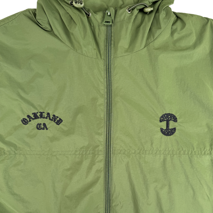 The Susurrus Track Top by Oaklandish is a light green nylon jacket with a classic fit, center zipper, and drawstring hood. "OAKLAND CA" is embroidered in black on the left chest, and a stylized tree appears on the right—an outerwear standout.