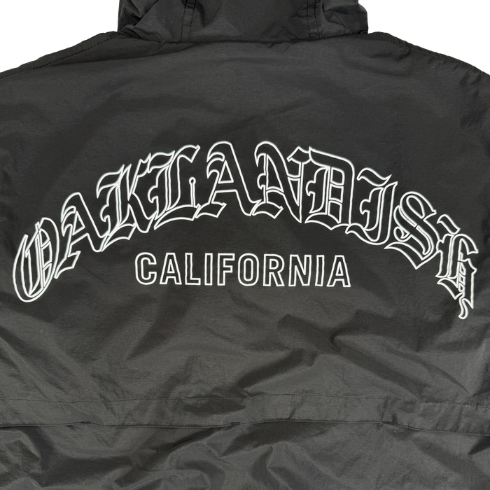 The Oaklandish Susurrus Track Top is a black men's zip jacket with bold white "OAKLANDISH" in gothic font above "CALIFORNIA" block letters on the upper back, shown laid flat to highlight the design.