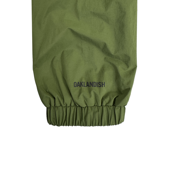 Close-up of the Susurrus Track Top by Oaklandish in olive green, showing the men's classic fit sleeve with an elastic cuff. "OAKLANDISH" is printed in black above the cuff, standing out against a plain white background.