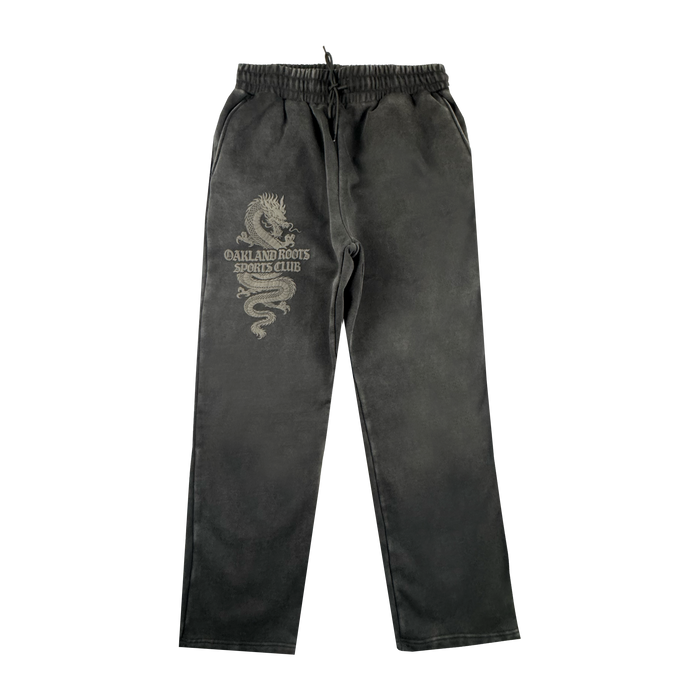 The Tatsu Pants by Oakland Roots SC are black organic cotton sweatpants with an elastic waistband, drawstring, side pockets, a relaxed straight-leg fit, and a light dragon graphic plus "OAKLAND ROOTS SPORTS CLUB" on the left thigh.