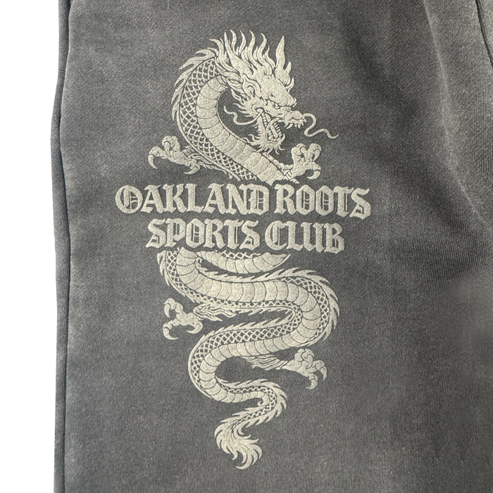 A detailed white dragon coils around bold, Gothic “OAKLAND ROOTS SC” text on dark gray organic cotton Tatsu Pants, capturing Oakland Roots pride.