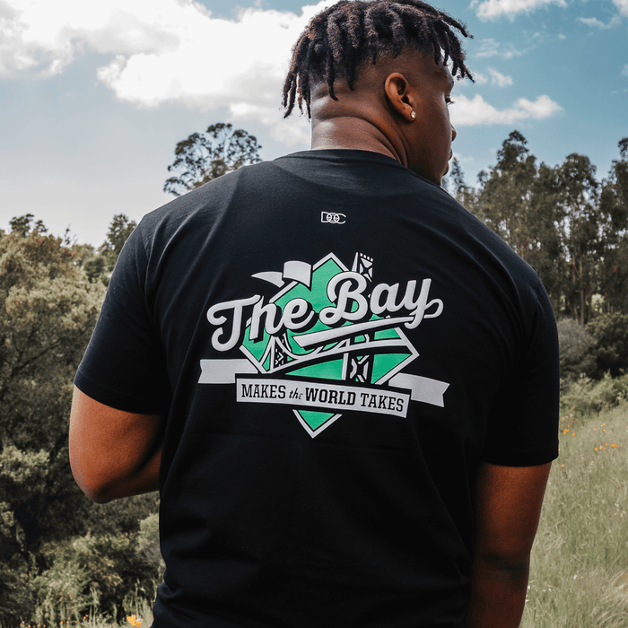 A person stands outdoors among trees and grass, facing away. They are wearing The Bay Tee by DOC from Other, a classic fit t-shirt featuring bold white and green text that reads “The Bay MAKES the WORLD TAKES” along with graphics of bridges and city icons—a true nod to Bay Area culture. The sky above is partly cloudy.