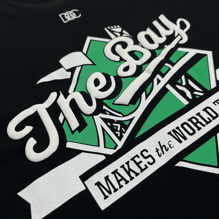 A close-up of The Bay Tee by DOC, part of The Bay Collection from Other, shows white text reading “The Bay” over a green geometric background, with smaller text below that says “MAKES the WORLD.” Inspired by Bay Area culture, the tee also features a small white “DSC” logo at the top of the design.