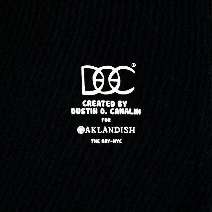 White text on a black background reads: “DOC Created by Dustin O. Canalin for Other The Bay–NYC,” with playful, stylized font for “DOC” and “Other.” The text is centered and clearly legible, capturing the spirit of Bay Area culture in this classic fit t-shirt, The Bay Tee by DOC from The Bay Collection.