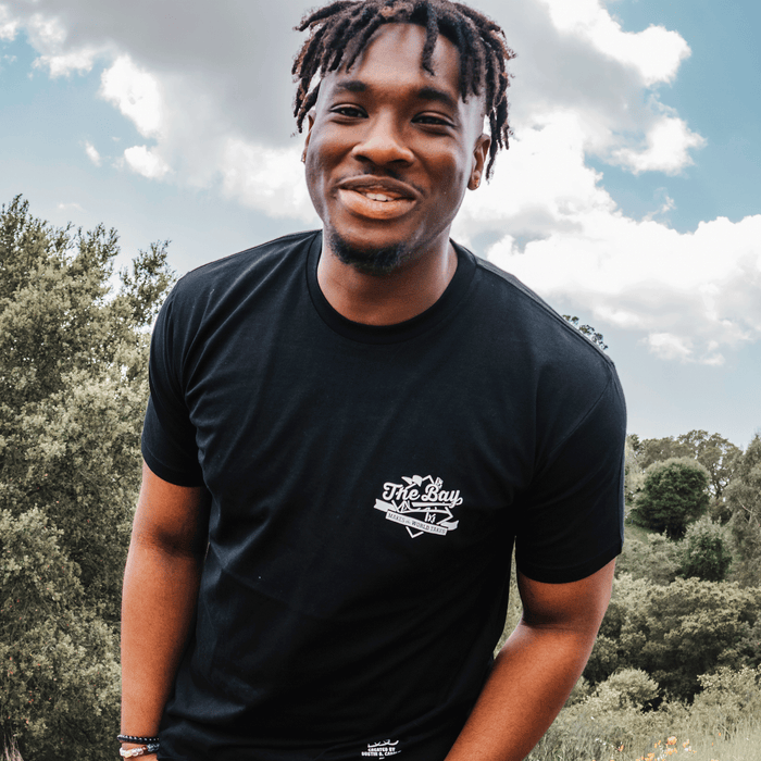 A young man with short dreadlocks smiles outdoors, wearing The Bay Tee by DOC from the brand Other. Trees and greenery fill the background under a sky with clouds. The bright, natural light gives the image a cheerful, relaxed feel that captures the essence of Bay Area culture.