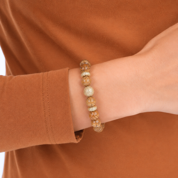 A close-up of a person's wrist wearing a beaded bracelet made of natural tan and brown wood beads with a textured silver-toned accent bead, set against a warm rust-brown long-sleeve shirt.