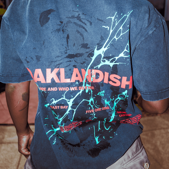 Wearing the Oaklandish Tiger Blood Tee, a vintage black, classic fit shirt with bold red "OAKLANDISH" text and turquoise paint splatter on the back. Made from heavy-weight cotton, it features partial “WHO WE DO IT” text. Photo taken from behind.