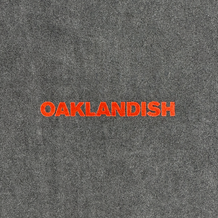 A close-up of the Tiger Blood Tee by Oaklandish shows gray, 100% cotton fabric with "OAKLANDISH" boldly embroidered in orange uppercase letters, centered on vintage black tee-like material against a plain background.