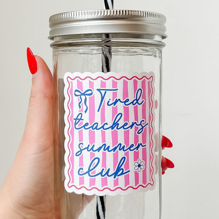 Hand holding mason jar tumbler up close that reads tired teachers club in blue ink with blue bow and flower on pink and white stripe background with red border