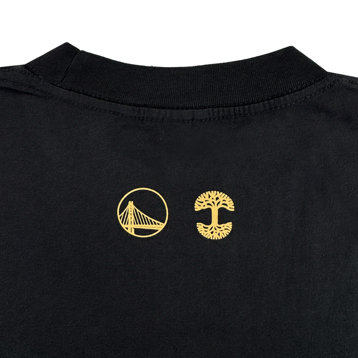 A close-up of the Town Doodle Tee by Other shows the back collar area featuring two yellow logos: a stylized bridge inside a circle on the left and a tree with visible roots and branches on the right. Both logos, part of the Warriors x Oaklandish Town Collection, are centered below the collar.