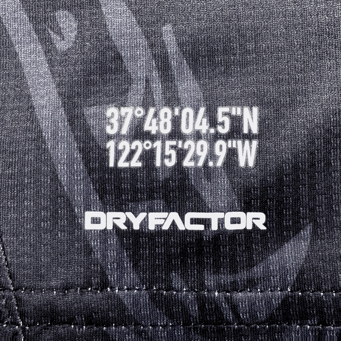 Close-up of dark textured fabric from the Unisex Oak-ness Awakens Kit by Oakland Roots SC. White coordinates "37°48'04.5"N 122°15'29.9"W" appear above bold "DRYFACTOR," with stitched lines and patterns inspired by Lake Merritt and Oakland folklore.