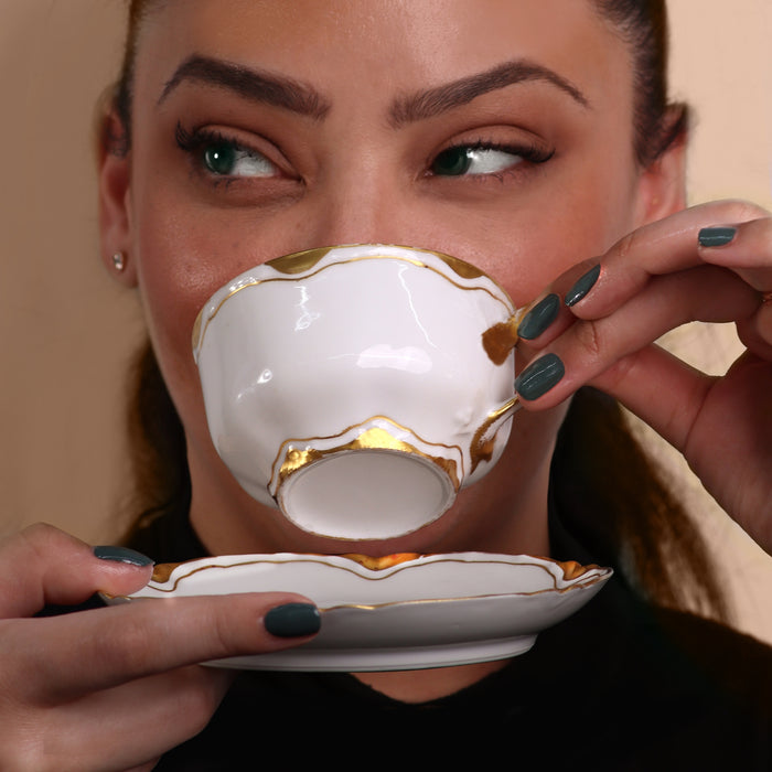 A person sips from a luxurious white and gold teacup with a matching saucer, their olive green LONDONTOWN Velvet Drapes nail polish adding a striking touch.
