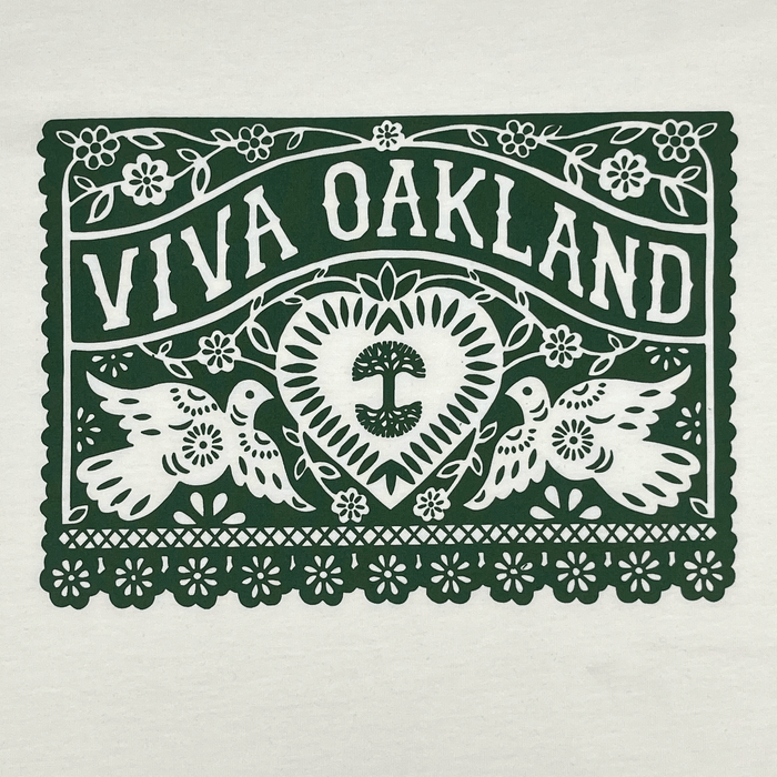 The Viva Oakland Tee by Oaklandish features a decorative design with the text "VIVA OAKLAND" in bold letters at the top. Below it, two birds flank a heart containing a tree emblem. The design includes intricate floral patterns and a scalloped border, all printed in dark green on a white background on this 100% cotton classic fit t-shirt.