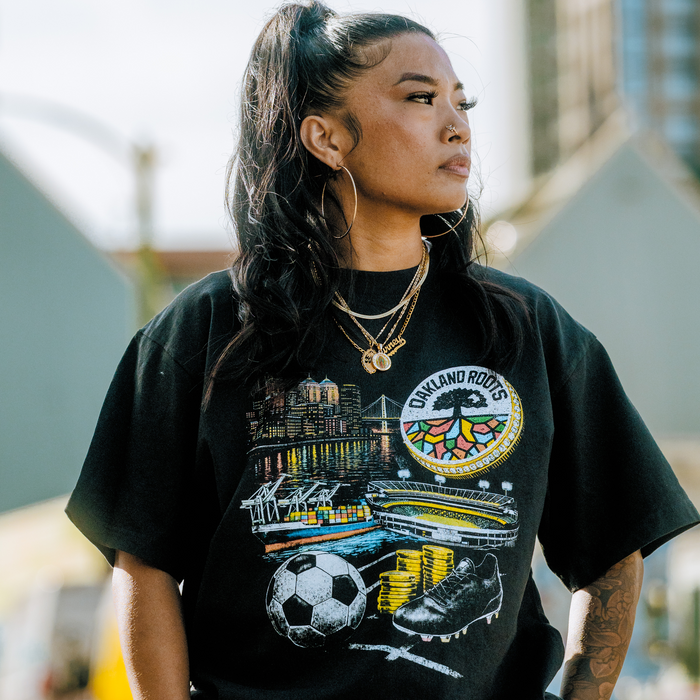 A woman with long dark hair in a high ponytail wears gold hoop earrings, layered necklaces, and the Oakland Roots SC Wager Tee with colorful graphics. She stands outdoors, looking to the side with city buildings blurred behind her.