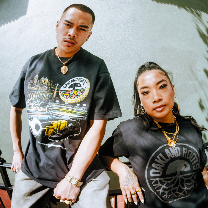 Two people stand confidently outdoors against a pale wall, both wearing Oakland Roots SC Wager Tees. The left features a scenic Oakland print; the right wears a black tee with the team's circular logo. Both look directly at the camera.