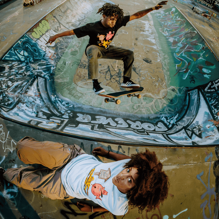 A skateboarder jumps over a person lying on a graffiti-covered skate park ramp. Both wear graphic tees featuring cartoon characters, with the skateboarder sporting the Wet Since ’88 Tee by Other, inspired by Oakland’s Digital Underground. The person on the ground looks up at the camera while the skateboarder is mid-air above them, frozen in action.