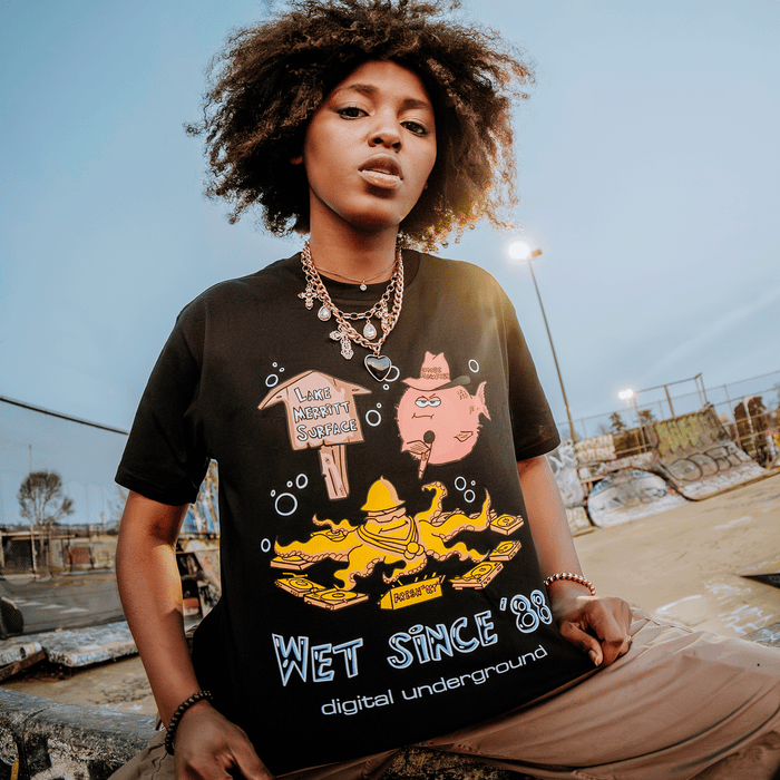 A young person with natural curly hair stands confidently at a graffiti-covered skatepark, sporting the “Wet Since ’88 Tee” by Other—a bold black graphic shirt adorned with cartoon characters, yellow rafts, and the bright “WET SINCE ’88 Digital Underground” text. Against an overcast sky that casts cool tones across the scene, their look perfectly channels an urban vibe reminiscent of Underwater Rimes.