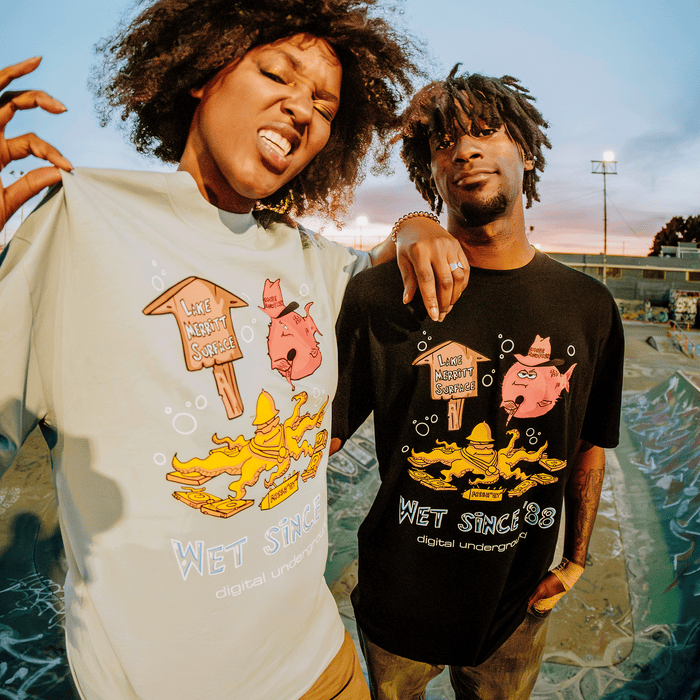 Two people pose at a skatepark in playful graphic tees from Other, each featuring cartoon fish, a wooden sign that reads “LIKE NOBODY SURFACES,” and “WET SINCE ’88”—the signature of the Wet Since ’88 Tee. One person mugs for the camera while the other looks serene, embodying Oakland t-shirt style and Underwater Rimes’ lively energy.