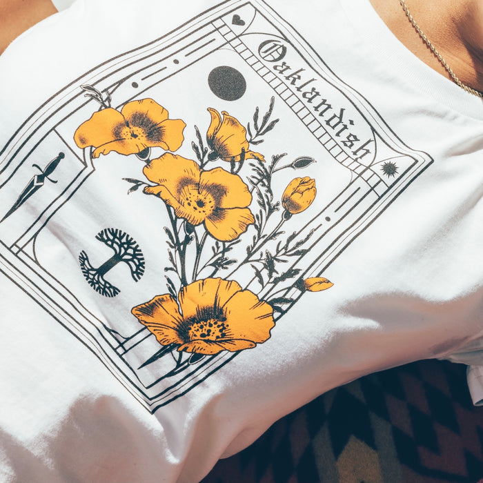 Close-up of a person wearing the Women's Blossom Tee from Oaklandish, a 100% cotton white t-shirt featuring an intricate design with yellow-orange poppies and botanical elements. The classic fit design resembles a vintage botanical print, with the word "Outlandish" at the top, bordered by decorative lines and small icons, including a crescent moon.