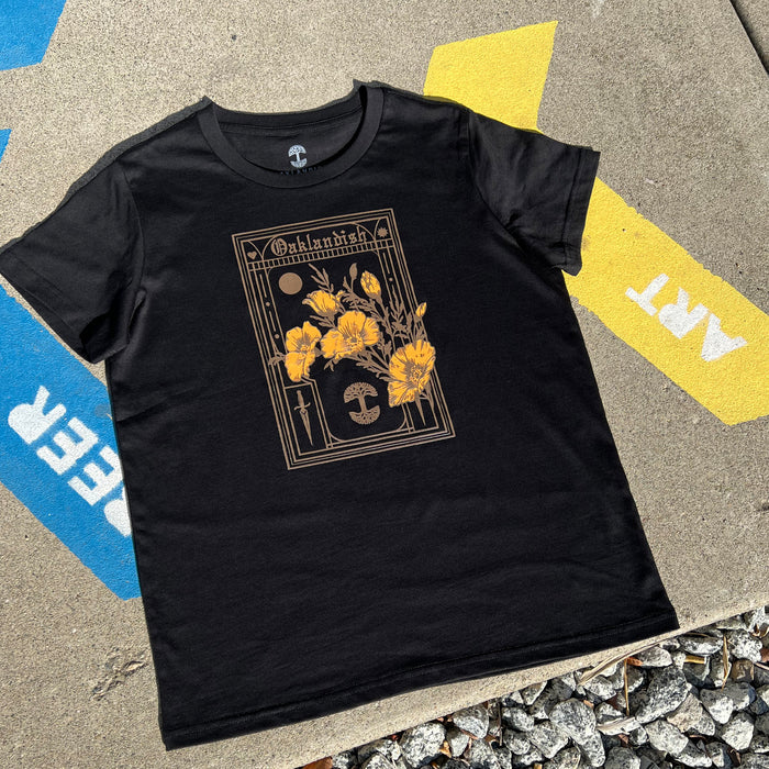 The Women's Blossom Tee by Oaklandish is a black T-shirt in women's sizing that showcases a detailed design on the front with yellow poppies, a moon, and the word "Oklahoma" framed ornately. The shirt is photographed laid flat on concrete pavement, partially over a yellow and blue painted section featuring the white text "FIRE" and "ART," with small rocks scattered at the bottom of the image.