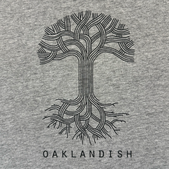 The Women's Oaklandish Logo Tee by Oaklandish features a stylized black tree with intertwining branches and roots centered on a classic fit grey 100% cotton shirt, with "OAKLANDISH" in bold capitals beneath the design.