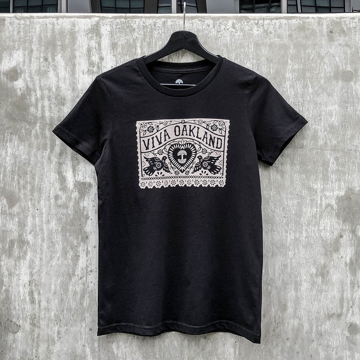 A black Women's Viva Oakland Tee from Oaklandish, made of 100% cotton, with a white design hangs on a black hanger against a concrete wall. The classic fit T-shirt features ornate floral patterns surrounding a central emblem with the text "VIVA OAKLAND" at the top and a stylized image of a heart with rays beneath it.