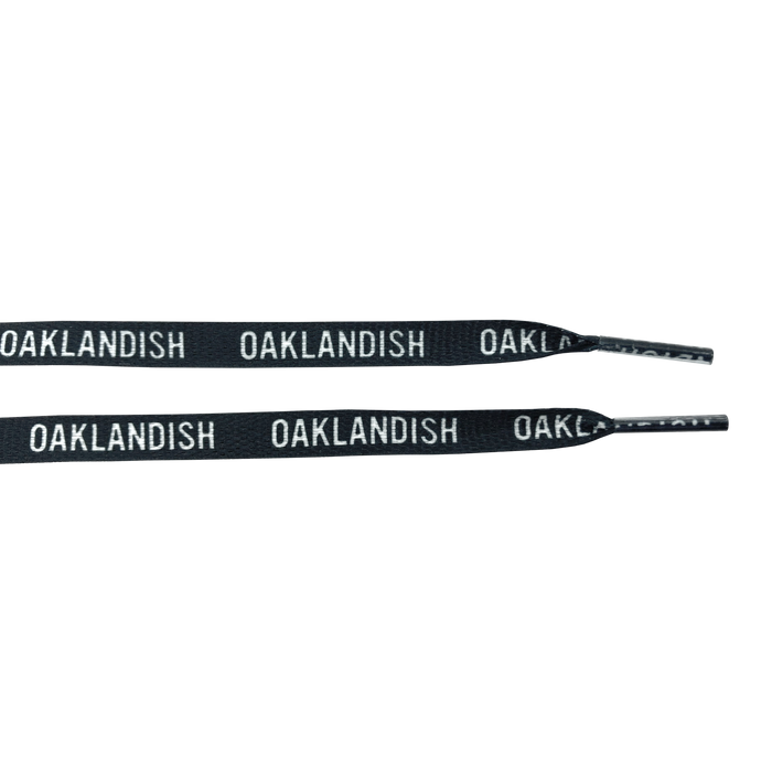 The "Wordmark Shoelaces" from Oaklandish are crafted from 100% black polyester and feature the word "OAKLANDISH" repeatedly printed in white, uppercase letters along their length. Resembling flat shoelaces, they are partially coiled to highlight the striking contrast between the black material and the white text, set against a plain white background.