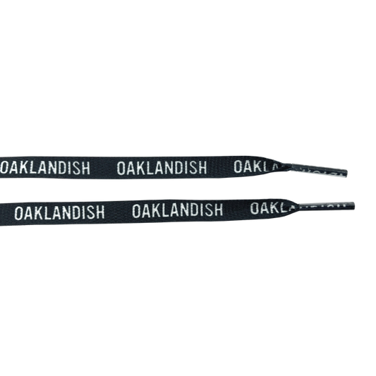 The "Wordmark Shoelaces" from Oaklandish are crafted from 100% black polyester and feature the word "OAKLANDISH" repeatedly printed in white, uppercase letters along their length. Resembling flat shoelaces, they are partially coiled to highlight the striking contrast between the black material and the white text, set against a plain white background.