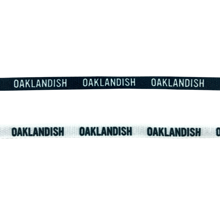 Two Wordmark Shoelaces are shown side by side. The top one is black with "OAKLANDISH" written repeatedly in white. The bottom one is gray with "OAKLANDISH" written repeatedly in black. Both 100% polyester shoelaces from Oaklandish have a similar design with the text spaced evenly along the length of each shoelace.