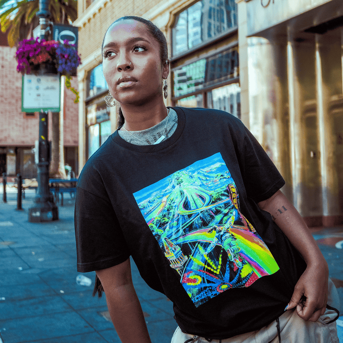 A person with long hair tied back stands on a city sidewalk in Oakland, wearing the Other Yay Area Tee—a black T-shirt featuring a colorful, abstract graphic. Buildings, palm trees, flowers, and street signs fill the bustling background in daylight, reflecting the vibrant energy often celebrated by painter and muralist portraiture specialists.