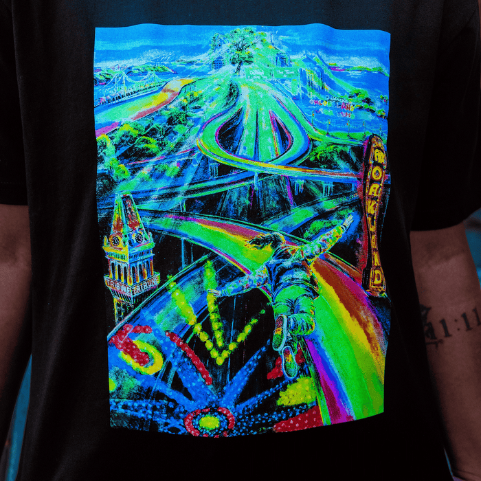 The Yay Area Tee by Other features a vibrant, psychedelic design channeling Oakland’s dynamic spirit—a flying figure soars above colorful, winding roads, abstract city buildings, and bold neon patterns. Reminiscent of muralist portraiture, “World” is displayed vertically in bright yellow on a sign. Blues, greens, yellows, and reds energize this surreal, futuristic scene.