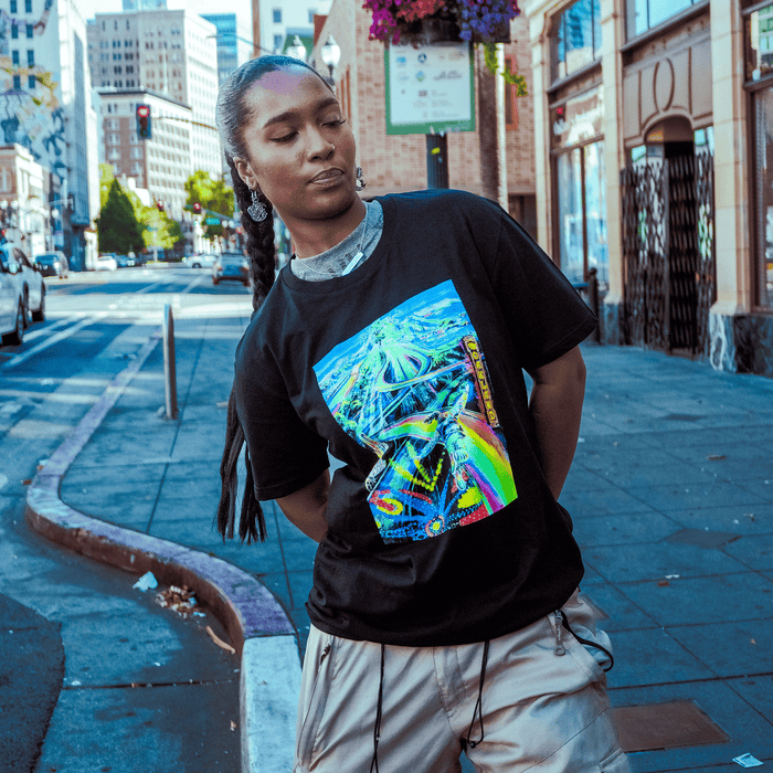 Sure! Here’s the revised sentence using your product data: A person with long braided hair stands on an Oakland city street, wearing the Yay Area Tee by Other—a black T-shirt featuring a colorful graphic—paired with beige shorts. Tall buildings, street signs, parked cars, and hanging flowers fill the background, evoking a vibrant scene of urban portraiture.