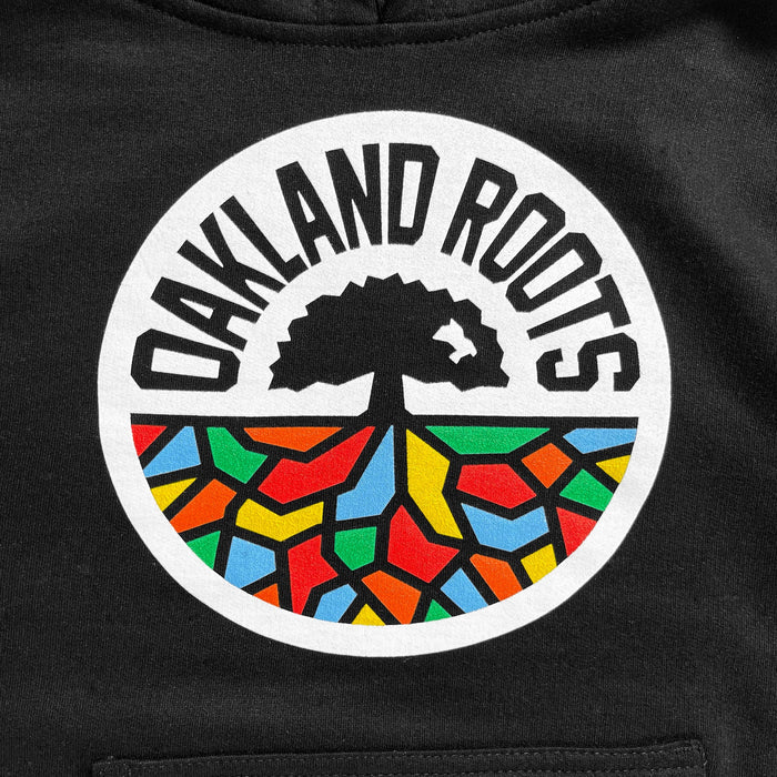 A Youth Oakland Roots SC Hoodie showcases a circular "Oakland Roots" logo on a black background. The logo features a black tree silhouette reminiscent of the Oaklandish design, positioned above colorful geometric shapes in red, yellow, green, and blue that resemble roots. These elements are all enclosed within a white circle with the text "OAKLAND ROOTS" arching over its top edge.